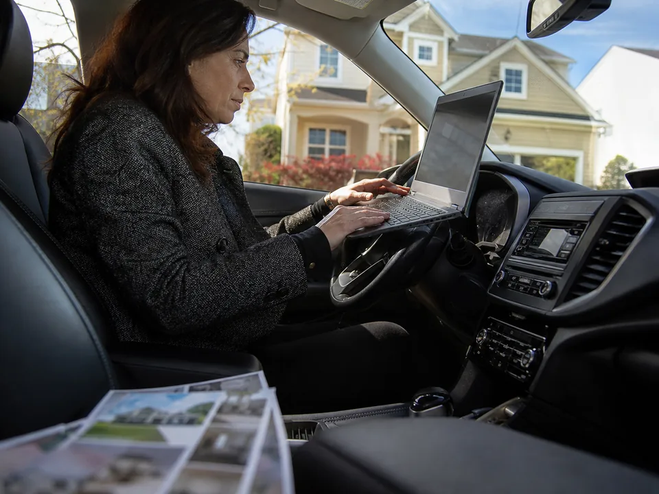 Real estate agent in her car between showings, drafting a listing post on a laptop